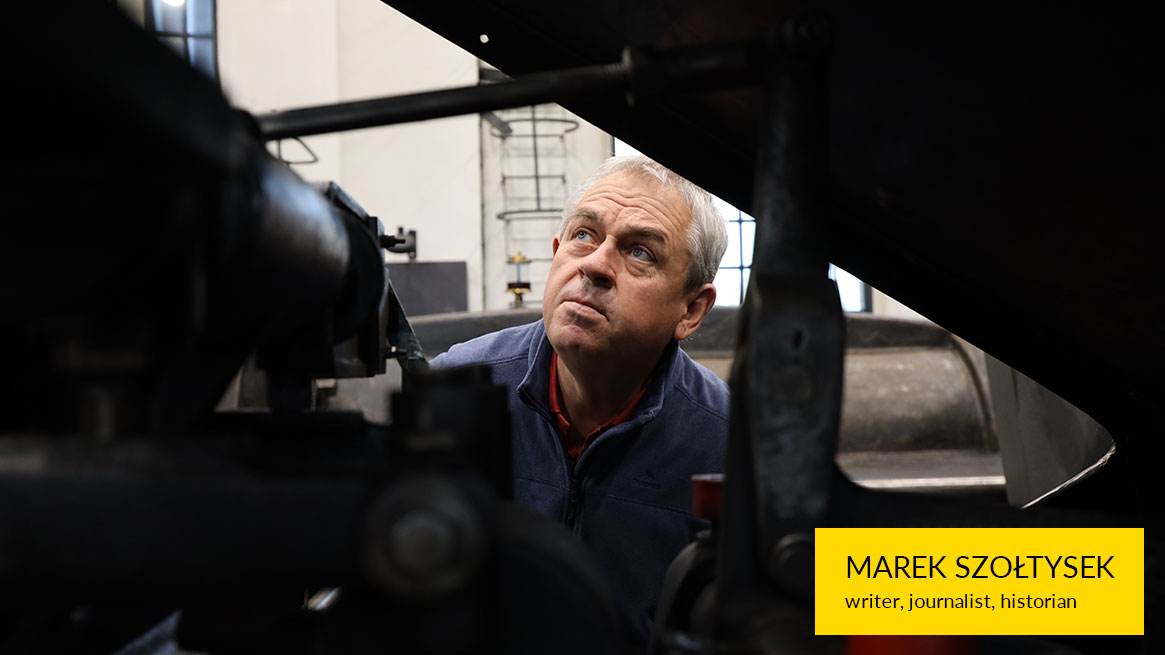 The photograph shows a man with short gray hair standing behind a machine. He is looking at it with great interest. The photo is taken through a crack in the device.