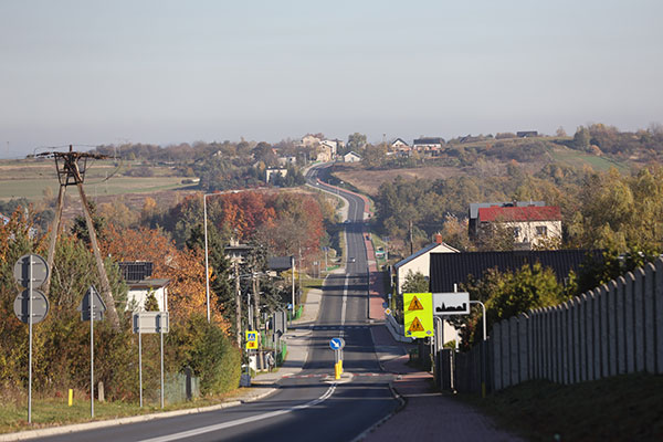 Na zdjęciu widzimy panoramę małej miejscowości, której osią jest odnowiona droga, po której poruszają się samochody. Po lewej stronie widzimy chodnik, po prawej zaś ścieżkę rowerową. Jest również odpowiednie oznakowanie pionowe i poziome.