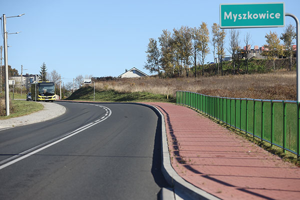 Zdjęcie wykonano wzdłuż odnowionej drogi. Po prawej widoczna jest ścieżka rowerowa oraz znak informujący o wjeździe do miejscowości Myszkowice. Z dala nadjeżdża autobus.