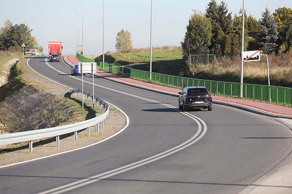 Na fotografii widoczna jest odnowiona jednopasmowa droga wyjazdowa z miejscowości, po której poruszają się samochody. Po lewej stronie znajdują się bariery energochłonne, po prawej ścieżka rowerowa i zielone ogrodzenie.