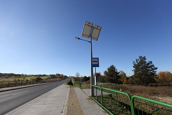 Fotografia przedstawia zatoczkę autobusową wzdłuż zielonej drogi. Na przystanku stoi latarnia, na której szczycie zamontowano panele fotowoltaiczne.