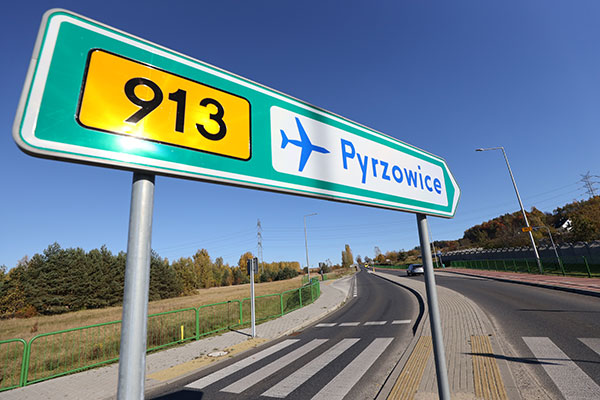 Zdjęcie przedstawia drogowskaz z napisem 913 Pyrzowice oraz symbolem samolotu. Ustawiony jest na wysepce oddzielającej dwa pasy ruchu o przeciwnym kierunku.