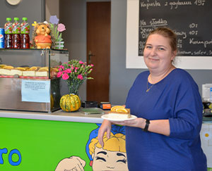 Zdjęcie ukazuje uśmiechniętą kobietę w niebieskim swetrze, która trzyma talerz z ciastkiem i opiera się o ladę.