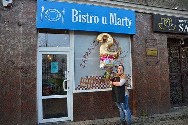 Fotografia przedstawia fasadę budynku z wejściem do bistro. Nad drzwiami widać napis „Bistro u Marty” na niebieskim tle. Przed wejściem stoi kobieta z balonami. Jeden z nich ma kształt cyfry dwa.