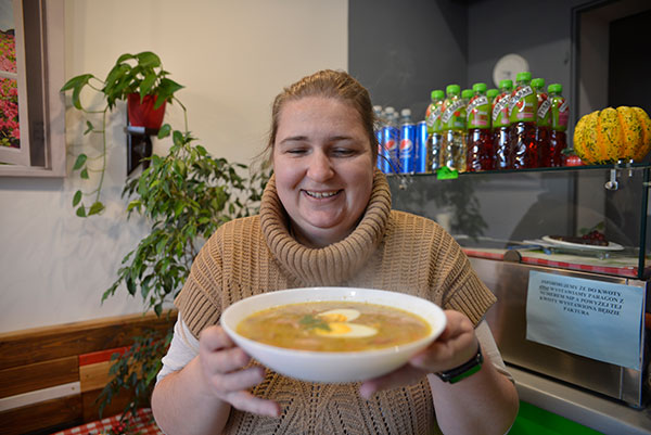 Na zdjęciu widzimy twarz uśmiechniętej kobiety, która unosi talerz parującej zupy.