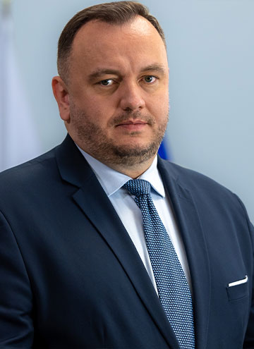 Fotografia przedstawia mężczyznę o ciemnych włosach, zaroście, w garniturze, pod krawatem. W tle widoczne są flagi Polski i Unii Europejskiej.