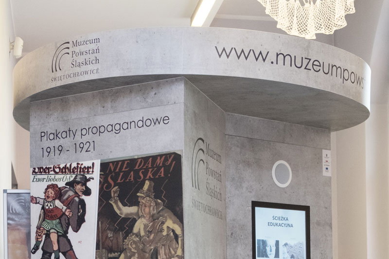 Fotografia przedstawia multimedialny słup używany w muzeum. Z widocznej strony znajdują się archiwalne plakaty propagandowe z czasów powstań.