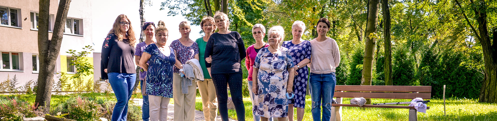 The photograph shows a group of women - seniors with their carers, standing by the pond. You can see a bench nearby and a tree and building in the background. The photo was taken by Dominik Wójcik.