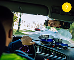 The photograph has been taken from the rear seat of the police car. You can see a mirror, which reflects the face of the policeman driving the car. Police hats are lying on the upholstery. The photo was taken by Maciej Motylewski.