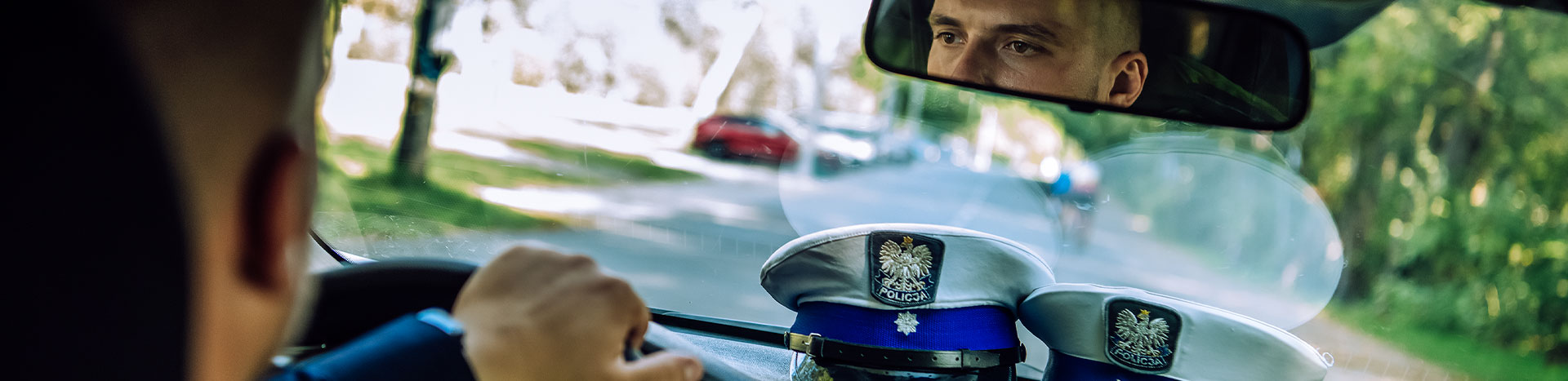 Fotografia jest wykonana z tylnego siedzenia radiowozu. Widać w nim lusterko, w którym odbija się twarz prowadzącego samochód policjanta. Na tapicerce widoczne są policyjne czapki. Autorem zdjęcia jest Maciej Motylewski.