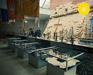 The photograph shows an exhibition in one of the rooms in Jasna Góra. This is a room where old brick wall elements are combined with new ones. The exhibited items are located in five glass showcases and are displayed on a shelf. You can see crosses, a typewriter, a model of a sailing ship, photographs and many other exhibits. The photo was taken by Dominik Wójcik.