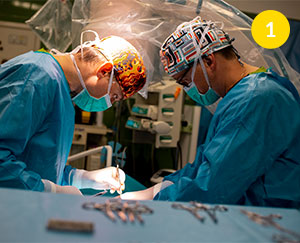 Fotografia przedstawia dwóch lekarzy weterynarii w czasie zabiegu na sali operacyjnej. Ubrani są w kolorowe czepki, maseczki i niebieskie fartuchy. Stoją naprzeciw siebie, a ich wzrok skierowany jest na stół operacyjny. Wokół widać instrumenty chirurgiczne. Autorką zdjęcia jest Lucyna Nenow.