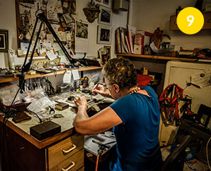 Fotografia przedstawia Małgorzatę Kupiec, która siedzi przy stanowisku pracy w pracowni i zajmuje się biżuterią. Widoczne są różne narzędzia, elementy biżuterii i kamieni. Poza tym, po prawej stronie segregatory, a nad stanowiskiem tablica korkowa z różnymi pocztówkami i notatkami. Autorem zdjęcia jest Dominik Wójcik.