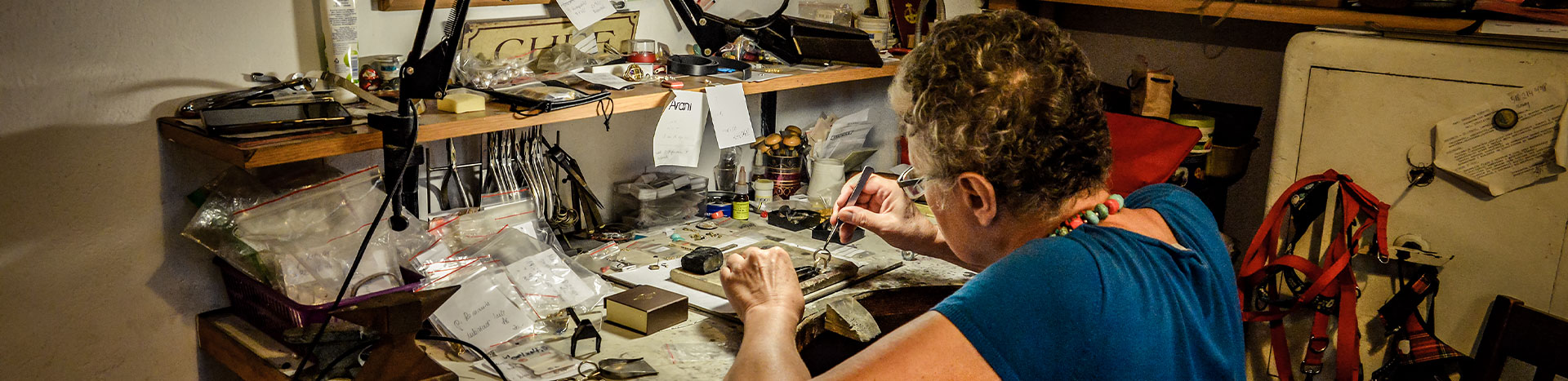 The photograph shows Małgorzata Kupiec, who is sitting at her workstation in the studio and working with her jewelry. You can see various tools, jewelry and stones. You can also see binders on the right side and a corkboard with various postcards and notes above the station. The photo was taken by Dominik Wójcik.