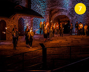 Fotografia wykonana jest na dziedzińcu zamku w Ogrodzieńcu. Przedstawia zwiedzanie nocne. Widać kilkanaście osób z pochodniami, które rozświetlają mrok i fragmenty zabytkowych murów, między innymi bramy. Zdjęcie pochodzi z archiwum beneficjenta.