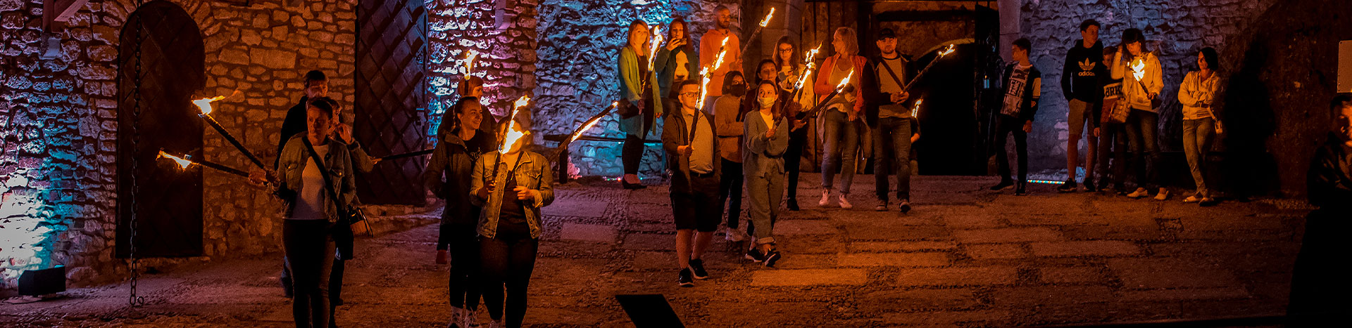 The photograph was taken in the courtyard of the castle in Ogrodzieniec. It presents a night tour. You can see a dozen or so people with torches that illuminate the darkness and fragments of historic walls, including the gates. The photo comes from the archives of the beneficiary.