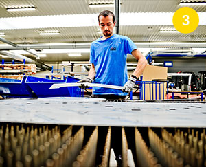 Fotografia przedstawia pracownika, który zbiera końcowy produkt pracy maszyny. To cienkie aluminiowe blachy, przycięte do odpowiednich kształtów. Autorem zdjęcia jest Dominik Wójcik.