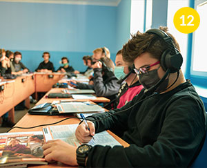Fotografia wykonana jest z poziomu stolika w klasie szkolnej. Widoczni są uczniowie ze słuchawkami na uszach, w trakcie zajęć językowych. Autorem zdjęcia jest Dominik Wójcik.
