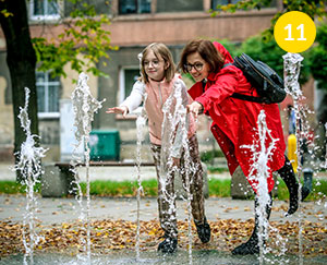 The photograph shows a woman in a red coat and her ten-year-old daughter. They are both wearing glasses. The are reaching out to the water gushing from the fountain. The photo was taken by Lucyna Nenow.