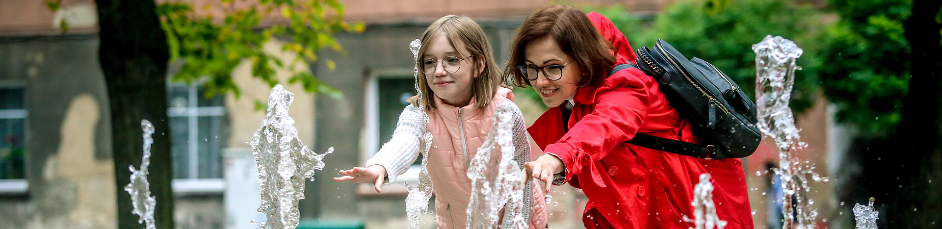 Fotografia przedstawia kobietę w czerwonym płaszczu oraz jej około dziesięcioletnią córkę. Obie w okularach, wyciągają ręce do wody tryskającej z fontanny. Autorką zdjęcia jest Lucyna Nenow.