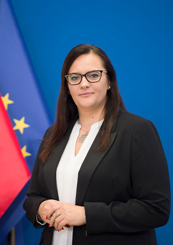 The photograph presents Małgorzata Jarosińska-Jedynak, the Secretary of State at the Ministry of Development Funds and Regional Policy. A woman has black, straight hair and she’s wearing glasses and a black jacket. She is standing against the background of the Polish and European Union flag. The photo comes from private archives.