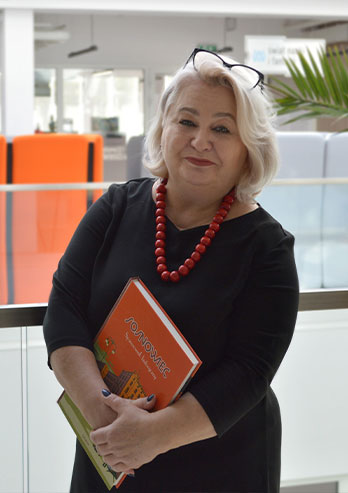 The photograph shows the director of the Zagłębie Mediateka, Elwira Kabat-Georgijewa. She has medium-length light hair. She’s wearing glasses on her head and red beads. She is standing inside Mediateka with a serene expression on her face. She's holding an orange book that says “Sosnowiec