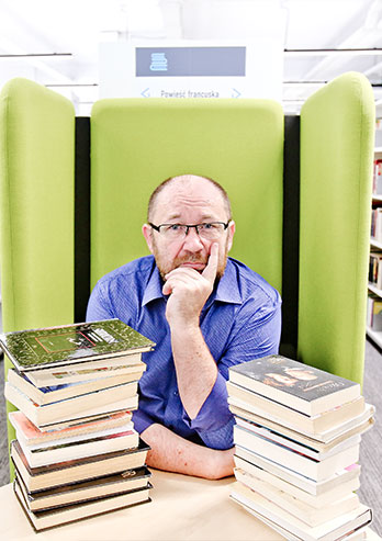 The photograph presents Professor Zbigniew Białas. The man is sitting at a table on a green armchair with a high backrest. There are bookcases on the right and left. You can also see books arranged in two piles lying on the table, at which the man is sitting. The professor has short hair, beard and glasses. He's looking towards the photographer. The photo was taken by Lucyna Nenow.