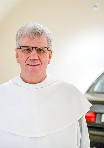 The photograph show a father Czesław Brud, administrator of the Pauline Monastery on Jasna Góra: A man with grey hair wearing glasses and a white cassock, with a cheerful expression on his face, is standing next to the car. The photo was taken by Maciej Motylewski.