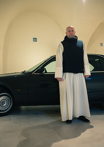 The photograph presents Father Wojciech Dec, the subprior of the Jasna Góra monastery. He is wearing a white cassock and a black vest and he’s standing next to the black BMW - one of the exhibits in the Jasna Gora Museum. The photo was taken by Dominik Wójcik.