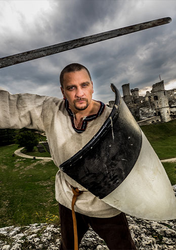 The photograph shows Wojciech Latacz, Eagle Nest trail guide showing visitors around the castle. The man is dressed as a knight and takes a combat pose. He has a black and white shield, a sword and clothes from the old days. In the background you can see the castle in Ogrodzieniec. The photo was taken by Tomasz Jodłowski.