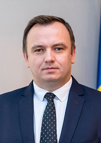 The photograph presents Jakub Chełstowski, the Marshal of the Silesian Voivodeship. The man is wearing a suit and a tie. We can see blue and yellow Silesian flag in the background. The photo comes from private archives.