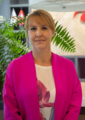 The photograph presents Anna Bobka Adwent, Director of the Upper Silesian Fund Branch in Katowice. The woman has light hair and a pink jacket. She’s smiling. Behind her there is a fern and a stand of the Upper Silesian Fund. The photo was taken by Maciej Motylewski.