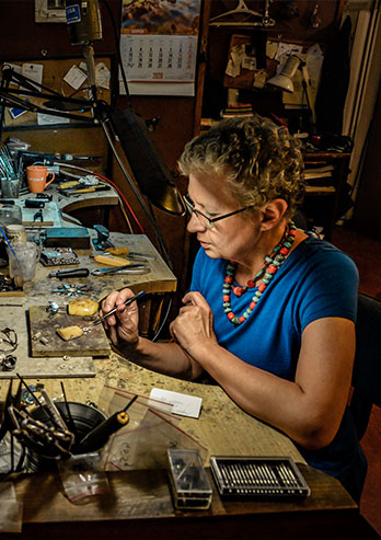 The photograph shows Małgorzata Kupiec, owner of a shop and a jewelry workshop. You can see her from the side. She’s working a piece of jewelry. You can see a lot of elements and tools. The whole room is dark, bright light from the lamp falls only on the stand and the face of the jewelry maker. The photo was taken by Dominik Wójcik.