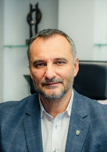 The photograph shows Krystian Grzesica, the mayor of Bieruń. He is a middle-aged man with short hair and a beard. He is wearing a shirt and a jacket. The photo was taken by Maciej Motylewski.