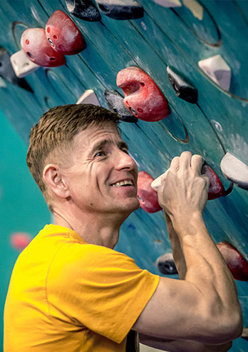 Fotografia przedstawia Sebastiana Wutke, wspinacza i trenera wspinaczki. Uśmiechnięty mężczyzna w żółtej koszulce wspina się na ściance. Autorką zdjęcia jest Lucyna Nenow.