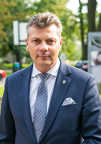The photo shows Mariusz Wołosz, the Mayor of Bytom. A middle-aged man is wearing a suit and a tie. He is standing against a walking path. The photo was taken by Maciej Motylewski.