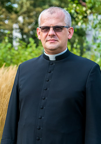 The photograph presents Father Krzysztof Karkoszka, parish priest of St. John Church in Rozbark, Bytom. The priest is a middle-aged man. He is wearing a cassock and glasses and you can see grain fields behind him. The photo was taken by Maciej Motylewski.