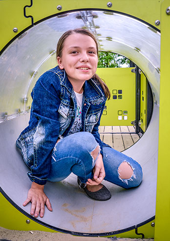 The photograph presents Marlena Sosnowska, a teenage participant of the day care center. The girl is wearing a denim jacket and she is sitting in a pipe, which is a part of the playground. She’s smiling. The photo was taken by Dominik Wójcik.
