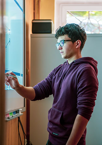 Fotografia przedstawia Marcina Sitko, ucznia Technicznych Zakładów Naukowych w Częstochowie. Młody chłopak w okularach i fioletowej bluzie, obsługuje interaktywną tablicę. Autorem zdjęcia jest Maciej Motylewski.