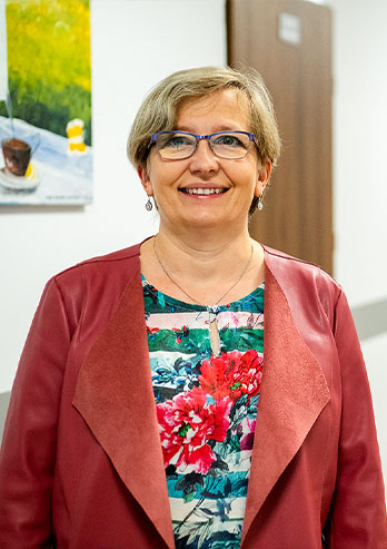 The photo shows Beata Bandura, Treasurer of the Board of The Thomas Apostle’s Hospice. A smiling woman in a red jacket and glasses is standing in the hallway of the Center. The photo was taken by Maciej Motylewski.