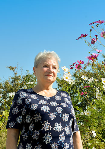 Fotografia przedstawia Beatę Walotek, podopieczną Centrum. Seniorka w granatowej sukience w kwiaty, stoi na tle kolorowych kwiatów. Autorem zdjęcia jest Dominik Wójcik.