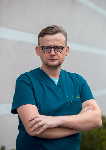 The photograph presents Oliver Teodorowski who has a title of PhD in veterinary sciences. The man is standing in front of the clinic building in a green short-sleeved uniform. His hair s combed to the side and he’s wearing glasses. He's looking straight ahead. The photo was taken by Lucyna Nenow.