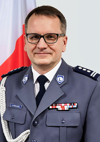The photograph shows the Deputy Chief of the Regional Police Headquarters in Katowice. A man in a ceremonial uniform is standing with a Polish flag in the background. He’s wearing glasses. He’s smiling. The photo comes from the archives of the beneficiary.