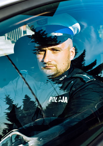 The photograph shows Kamil Adamczyk, a junior warrant officer. A policeman is sitting in a police car, in the driver's seat. He's looking towards the photographer from behind the window. The photo was taken by Dominik Wójcik.