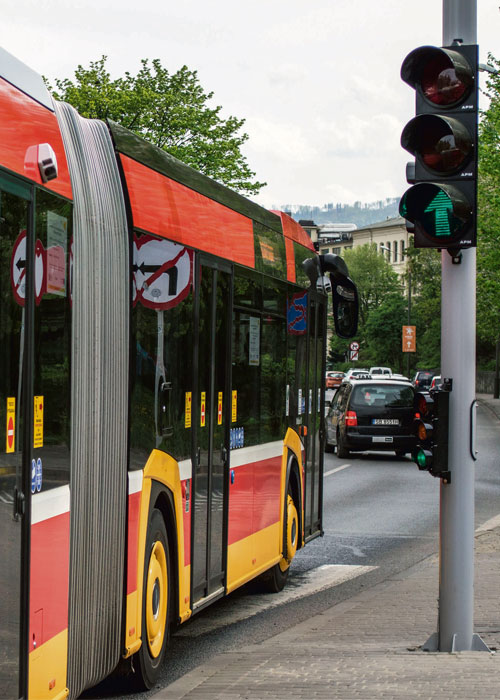 autobus w ruchu miejskim stoi przy sygnalizatorze wyświetlającym sygnał zielony. Przed nim samochody. W tle zieleń miejska i budynki.