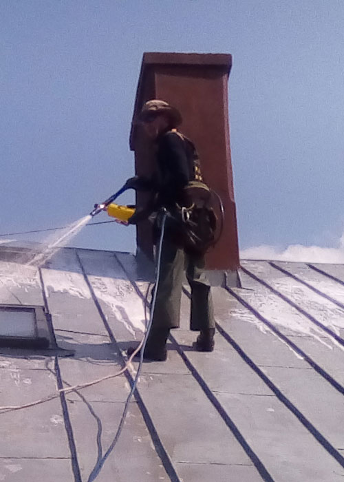 Mężczyzna maluje dach wyposażony w profesjonalny sprzęt. Ubrany jest w  strój roboczy i zabezpieczony liną. W dachu widoczne są dwa kominy i trzy okna.