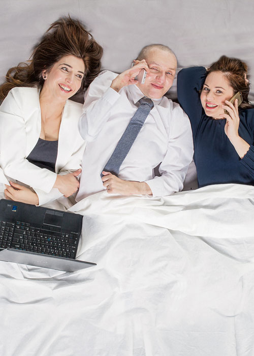 Trzy młode osób leżą obok siebie na łóżku, do połowy przykryci kołdrą. Ubrani elegancko w garnitury i garsonki. Śmieją się, rozmawiają przez telefon. Przed nimi znajduje się otwarty laptop.
