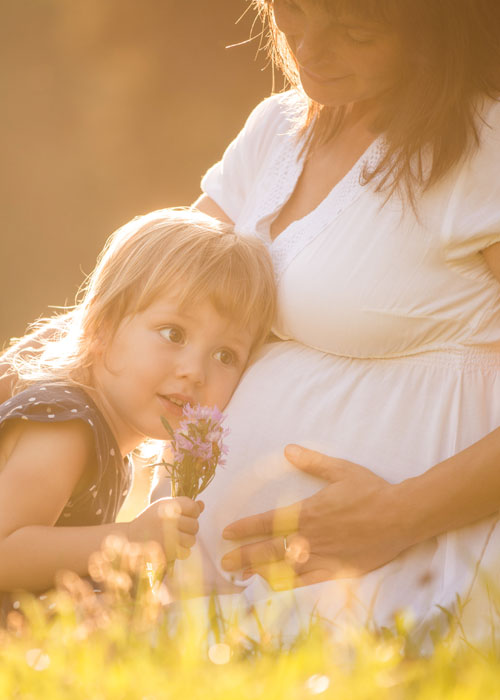 Na łące w słoneczny dzień kobieta w ciąży w białej letniej sukience  siedzi na trawie i przytula do brzucha małą dziewczynkę, która trzyma w ręce kwiaty.